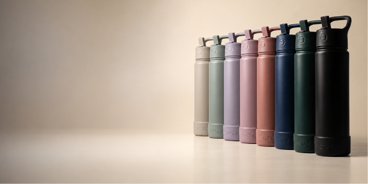 A lineup of eight  DOZZE Monochrome Edition water bottles in assorted colors, including almond beige, sage green, Mauve purple, creamy pink, guava orange, ocean blue, sacramento Green, midnight black  arranged on a light beige surface against a matching beige background.